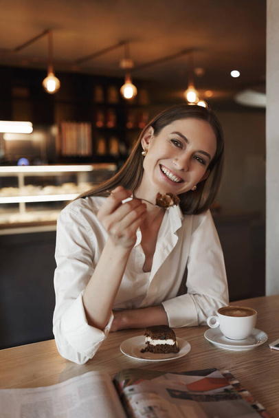 Treat yourself. Charming business woman, office lady in cafe, eating delicious dessert, holding fork with cake and drink coffee, reading interesting magazine, smiling happy, have chat with friend - Foto, afbeelding