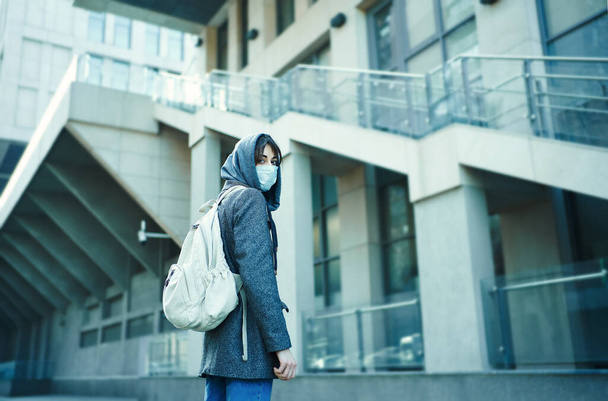 woman in hood with backpack turning around, standing against modern building in face mask for protection of coronavirus. - 写真・画像