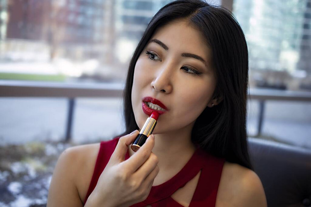 beautiful young Chinese Asian female model is wearing a fashionable sexy red dress. she begins to apply red cosmetic lipstick sensually to her lips - Foto, Imagen