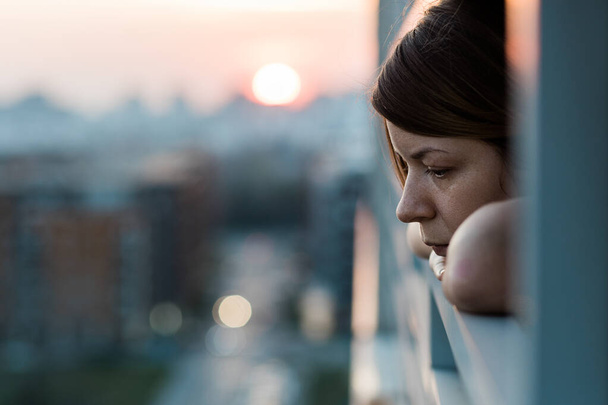 Joven mujer triste mirando hacia fuera a través del balcón de un edificio de apartamentos
 - Foto, Imagen
