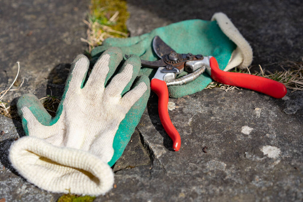 Πράσινα κίτρινα λαστιχένια γάντια με κόκκινα ανοιχτά κλαδευτήρια στο δρόμο κήπο γρανίτη στον ήλιο. Εργαλεία κήπου σε χρήση, προβολή υψηλής γωνίας. - Φωτογραφία, εικόνα