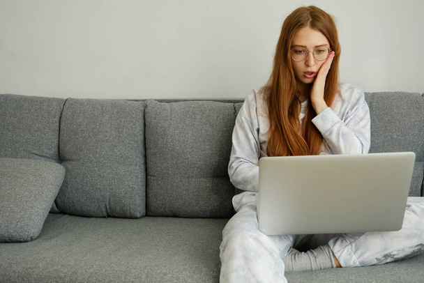 A red-haired girl with glasses, with long hair isits on the couch in a half-turn and looks intently at the laptop, opening mouth - Foto, immagini