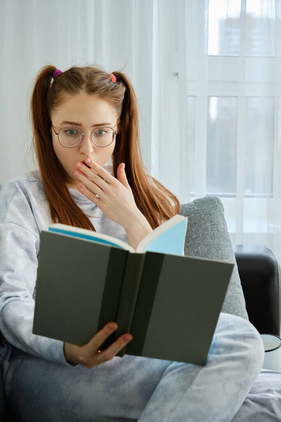 A redhead girl with glasses, with long tails in pajamas sits on the couch and looks in shock at the book, covering mouth with hand - 写真・画像