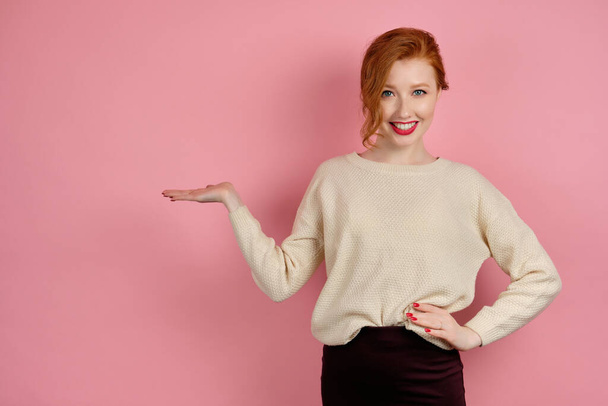 Cute red-haired girl with red lipstick and collected hair smiles on a pink background pointing her palm to the side - Photo, Image