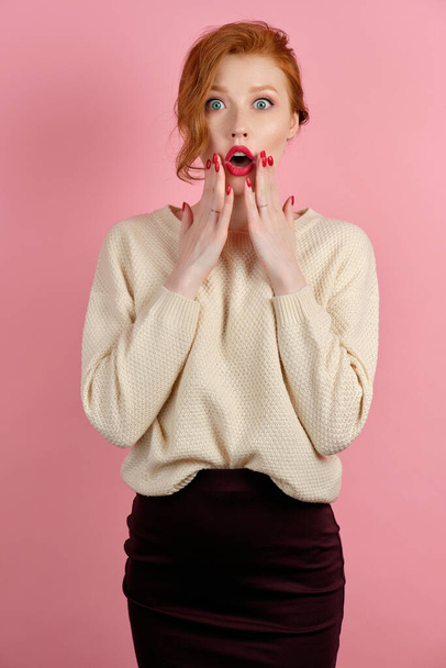 Redhead girl with red lipstick and red manicure looks shocked at the camera, raising her hands to her face - Foto, Bild