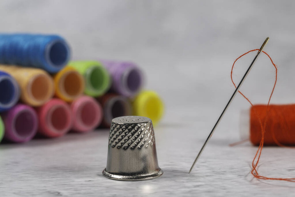 Visão de close-up de uma agulha com fio vermelho e dedal com bobinas de fio de cor diferente no fundo
 - Foto, Imagem
