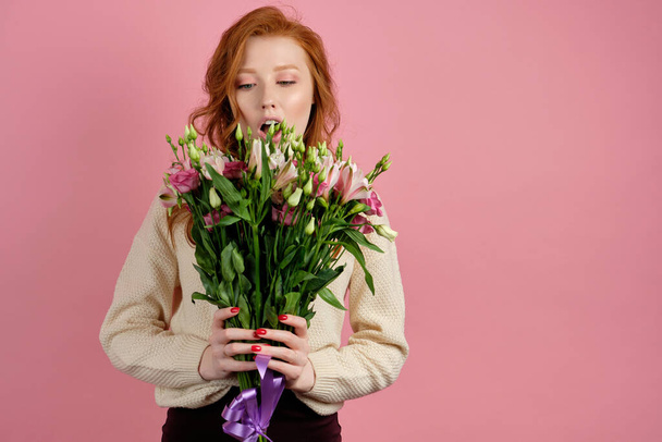 A beautiful red-haired girl stands on a pink background and looks at a large bouquet in her hands, opening mouth - Photo, image
