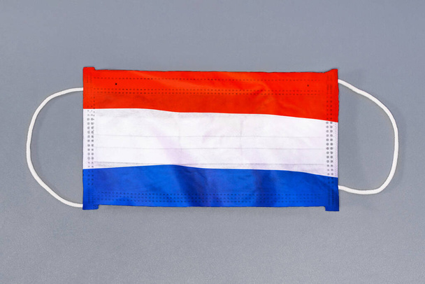 Gri arka planda Hollanda bayrağı olan izole tıbbi maske. Yakından koruyucu maskeler tekstil filtresi. Sağlık ve tıbbi konsept. Coronavirus, Hollanda 'da virüs.. - Fotoğraf, Görsel