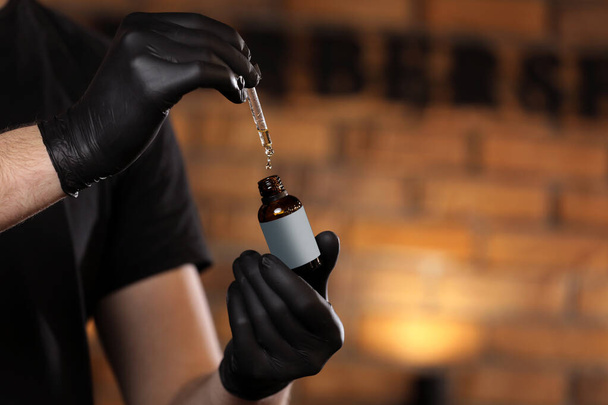 Man holding essential oil for beard or aromatherapy in glass bottle on hand and dripping oil. man with black gloves in barber shop. copy space on bottle. - Photo, Image