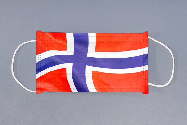 Μεμονωμένη ιατρική μάσκα με σημαία της Νορβηγίας σε γκρι φόντο. Στενές προστατευτικές μάσκες υφασμάτινο φίλτρο. Ιατρική περίθαλψη και ιατρική έννοια. Ιός του Coronavirus στη Νορβηγία. - Φωτογραφία, εικόνα
