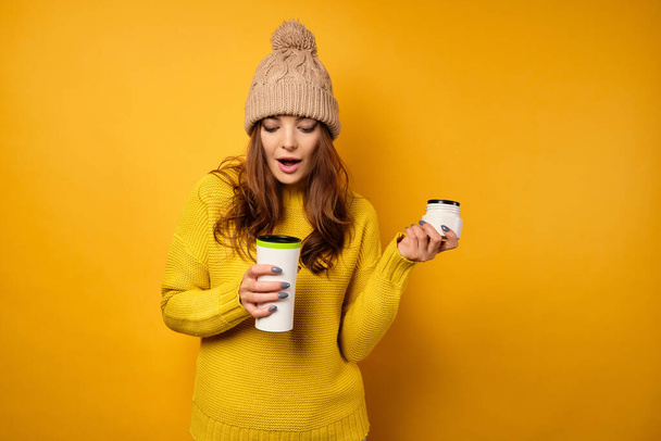 A beautiful brunette in a yellow sweater and hat stands on a yellow background, looks in a thermos, opening her mouth - Фото, изображение