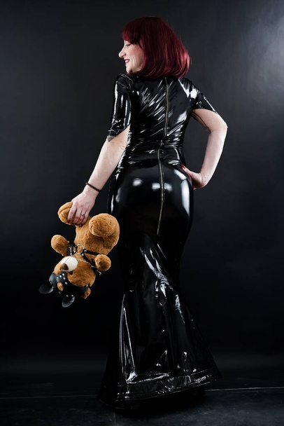 Strong BDSM woman in long fetish latex dress posing with bad bear in leather harness on dark studio background alone - Foto, imagen