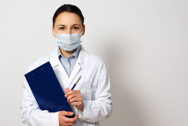 portrait of a female doctor in a protective mask with a clipboard and a pen in her hands, making a record. on white background. - Photo, image