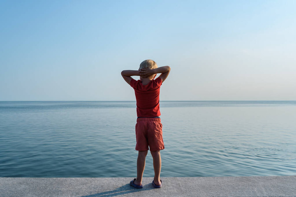 Dítě stojí na pobřeží a dívá se do dálky s rukama za hlavou. Pohled zezadu, na obloze a na moři. - Fotografie, Obrázek