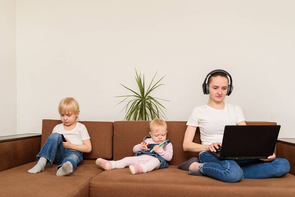 Mladá matka a dvě děti sedí na gauči s počítačem a telefonem. lidé a technologická závislost. - Fotografie, Obrázek