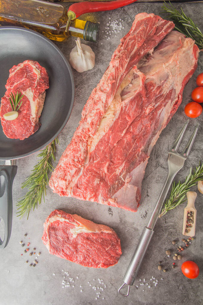 Een groot stuk rundvlees gesneden in steaks, waarvan er één al in een pan zit met kruiden. Raw New Yorkse biefstuk, bovenaanzicht. - Foto, afbeelding