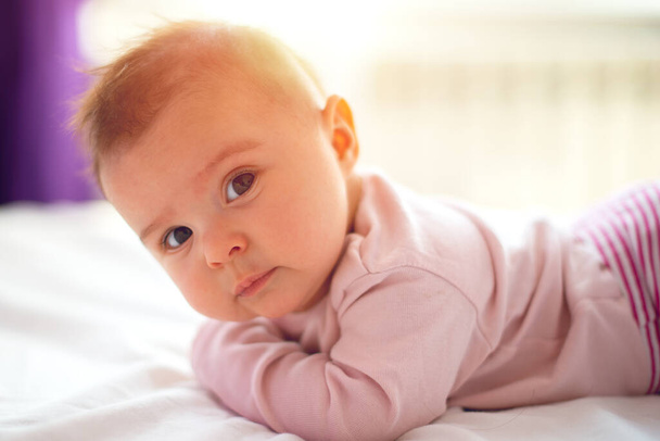 Cute three months old Baby girl infant on a bed on her belly with head up looking into camera with her big eyes. Natural bedroom light. - Photo, Image