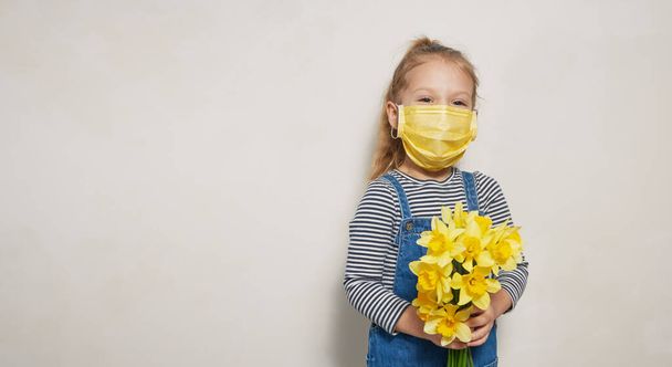 Petite fille enfant dans un masque médical de protection tient un bouquet de fleurs jaunes de printemps. Concept de quarantaine mondiale pandémique
 - Photo, image
