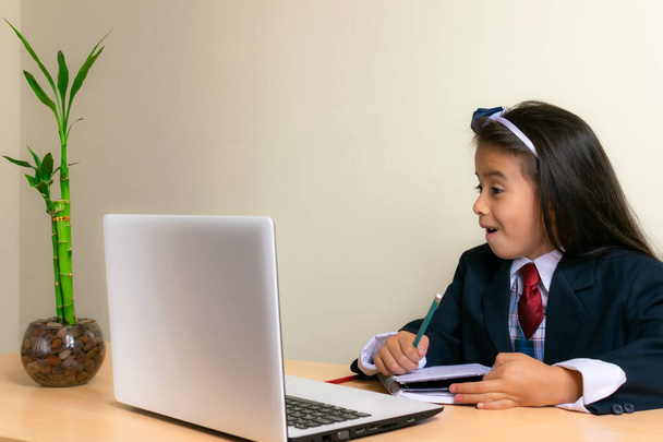 прекрасна колумбійська дівчинка, яка сидить за столом у класі, здивована, виконуючи домашнє завдання на комп "ютері. - Фото, зображення