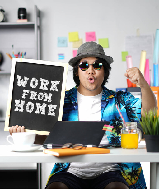Young Man Holding Work From Home Text on Blackboard While Working on Summer Vacation Season at Home - Photo, Image