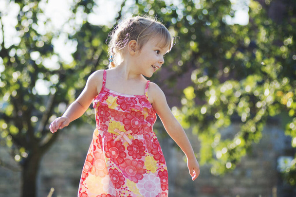 Schattig klein twee jaar oud meisje in rood bloemen jurk is buiten. - Foto, afbeelding