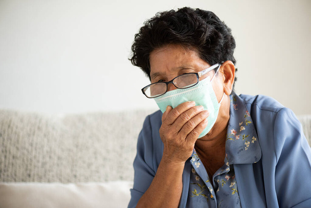 保護顔のマスクを身に着けている病気のアジアの老婦人は、自宅でソファに座って、コロナウイルスと午後2時半の戦いの準備をしてください。シニアヘルスケアコンセプト. - 写真・画像