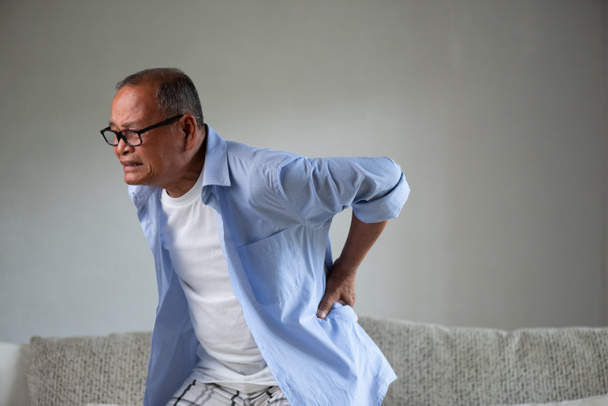 Asiatischer alter Mann sitzt auf dem Sofa und hat Rückenschmerzen, Rückenschmerzen zu Hause. Seniorenpflegekonzept. - Foto, Bild
