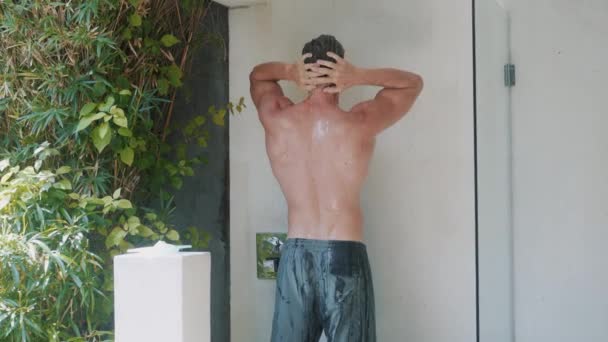 backside view of handsome man with strong body takes shower in villa cabin - Filmati, video