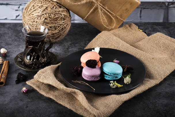 Bunte Macarons in einer schwarzen Untertasse und ein Glas Tee auf einem schwarzen Tisch - Foto, Bild