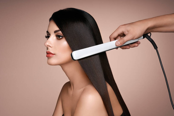 Hairdresser straightening long dark hair with hair irons. Beautiful woman with long straight hair. Smooth hairstyle - Foto, Imagem