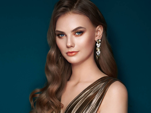 Brunette girl with perfect makeup. Beautiful model woman with curly hairstyle. Care and beauty hair products. Lady with fashionable gold makeup. Model with jewelry on dark blue background - Photo, Image