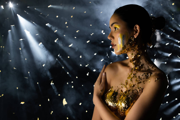 Fashion Young Asian Woman eyes black wrapped hair beautiful make up fashion decorate with Golden Foil or Gold leaf all over chest. Studio Lighting dark Background star holes smoke - 写真・画像
