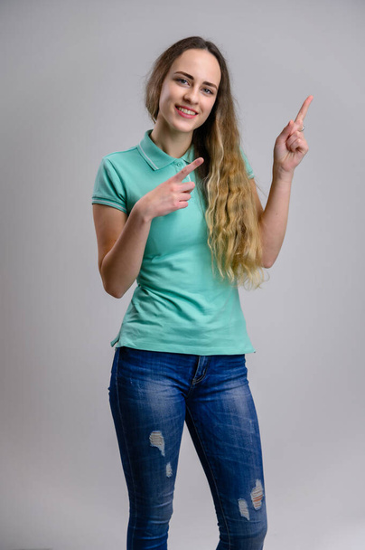 Vertical photo of a pretty young woman with long hair in a turquoise blue jeans blouse on a white background. Model points fingers to the sides. - Photo, image