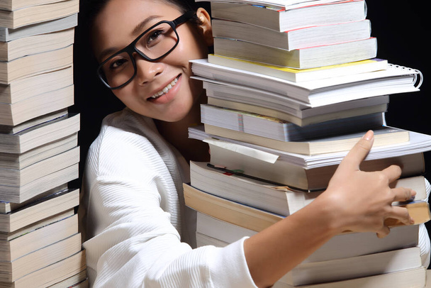 Mooi aziatisch meisje glimlach en blij om tijdschrift te lezen tussen stapelen van vele soorten boeken, leerboeken, tijdschrift, verslag, onderzoek, strip in twee torens op tafel met donkere zwarte achtergrond nacht tijd - Foto, afbeelding