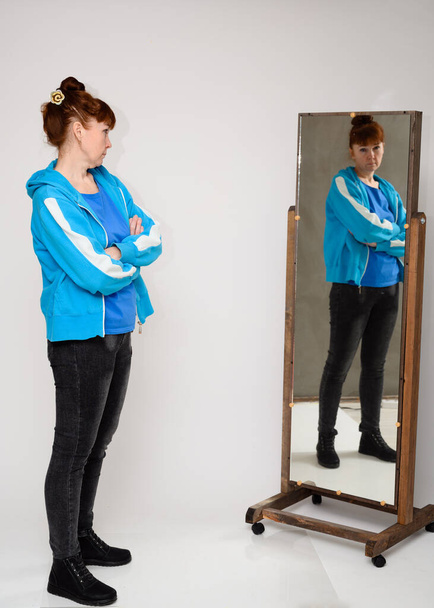 Vertical photo of a woman in a blue blouse and jeans standing in front of a mirror on a white background - Foto, afbeelding