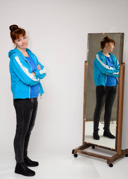 Vertical photo of a woman in a blue blouse and jeans standing in front of a mirror on a white background - 写真・画像