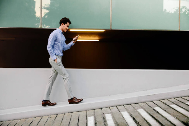 Feliz joven hombre de negocios en ropa casual usando el teléfono móvil mientras camina por el muro del edificio urbano. Lifestyle of Modern People. Vista lateral. Longitud completa
 - Foto, imagen