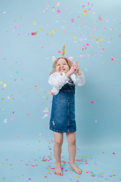 Happy little blonde girl in unicorn head band and jeans overalls celebrating, clapping her hands over blue background. - Valokuva, kuva