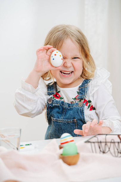 Happy playful little girl sitting behind a table with easter eggs, holding one over her eye. Looking at camera. Grimasing, making scary faces. - Фото, изображение