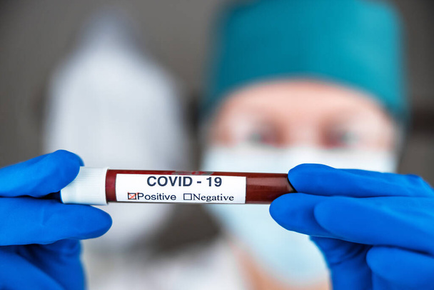 A nurse in a respiratory mask holds a test tube labeled COVID 19, with a positive blood test for a new rapidly spreading coronavirus, close-up, shallow depth of field, selective focus. The concept of - Fotó, kép