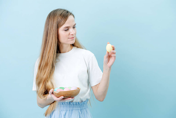 Happy pleased woman standing with a bowl of easter eggs in hands over blue background. Holding one egg in her hand, looking at it. She has an extra long beautiful hair and casual clothes. - Foto, Imagem
