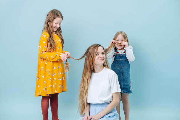Two little girls playing with their mother's hair over blue background. One making braid, while second one making hair mustache and laughing. Happy mom looks at camera. - Valokuva, kuva