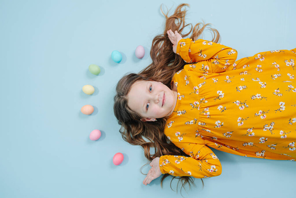 Happy little brunette girl in orange shirt lies on a blue-surfaced floor with a rainbow of easter eggs over her head. Top view. - Foto, immagini