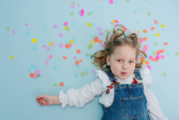 Happy little blonde birthday girl in jeans overalls lies on a blue-surfaced floor with colorful confetti pieces over her head. Top view. Inflating fer cheeks, making funny cute face. - Foto, Imagen