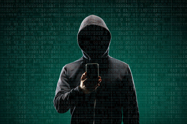 Dangerous hacker with a smartphone gadget over digital background with binary code. Obscured dark face in mask and hood. Data thief, internet attack, darknet fraud, virtual reality and cyber security - Photo, Image