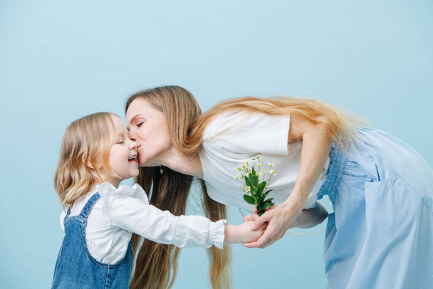 Little girl gives her mother bouquet of wild camomiles, she kisses her for it. Both look happy. Over blue background. - Photo, Image