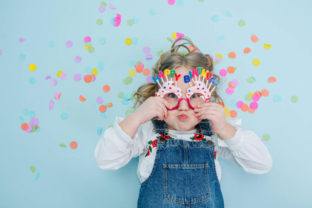 Happy little blonde girl in happy birthday glasses jeans overalls lies on a blue-surfaced floor with colorful confetti pieces over her head. Top view. Inflating fer cheeks, making funny face. - Foto, imagen