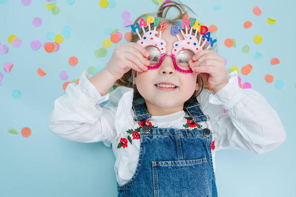 Happy little blonde girl pressing happy birthday glasses to her face, wearing jeans overalls lies on a blue-surfaced floor with colorful confetti pieces over her head. Top view. Funny face. - Zdjęcie, obraz