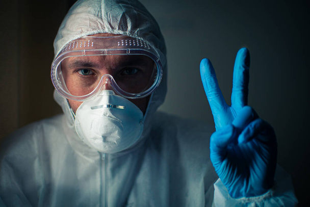 呼吸器、眼鏡、細菌学的保護スーツの疲れている表情の医者は青い医療用手袋で手で勝利の兆候を示しています。医療従事者への献身の概念 - 写真・画像