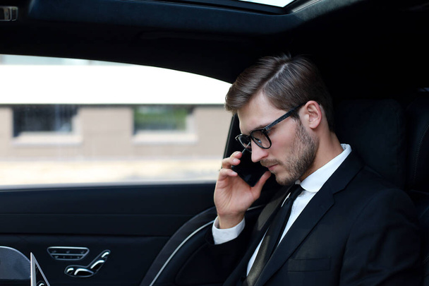 Handsome businessman talking with phone sitting with laptop on the backseat of the car - 写真・画像